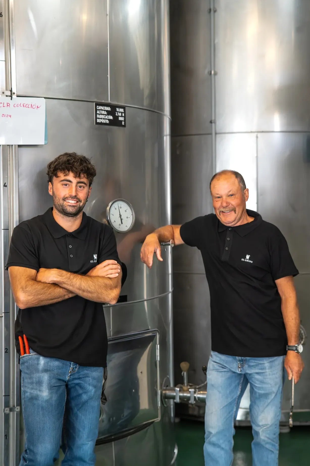 Dos trabajadores de la bodega posan sonrientes frente a los depósitos de acero inoxidable en la zona de elaboración, reflejando profesionalidad y cercanía.
