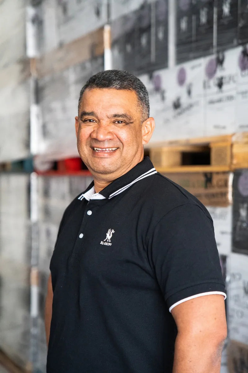 Hombre sonriente con camiseta negra tipo polo, de pie en un almacén con estanterías y cajas apiladas al fondo.