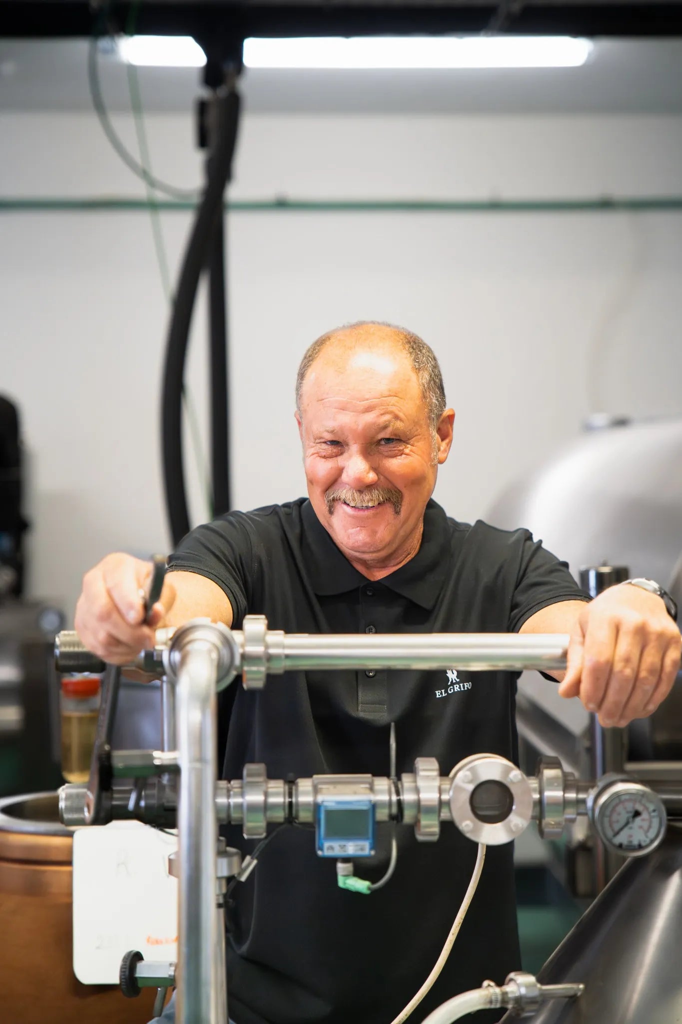 Hombre mayor sonriente operando una máquina industrial de acero inoxidable en una bodega, con tuberías y manómetros, vistiendo un polo negro con el logotipo de El Grifo